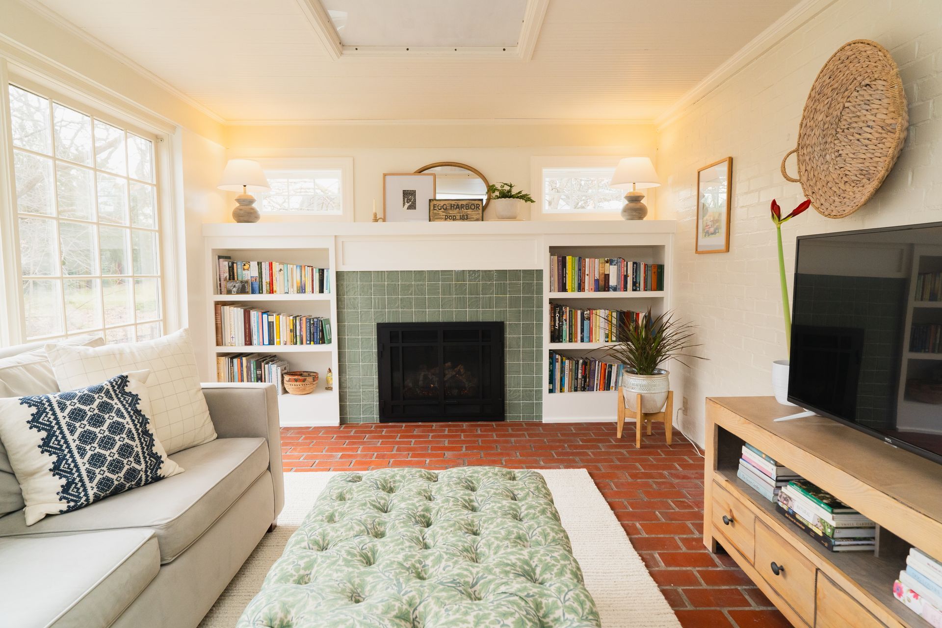 Bright living room with brick fireplace, bookshelves, beige sofa, green rug, and TV console