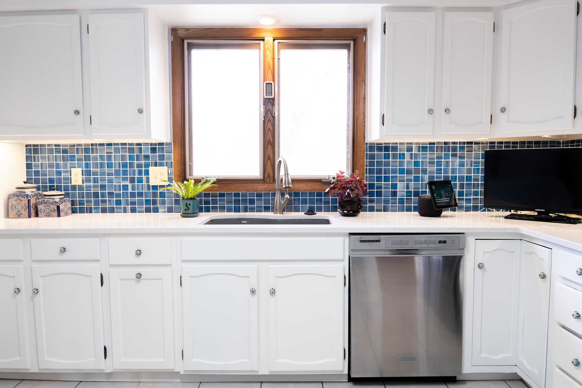 Bright white kitchen with blue tile backsplash, wooden window, sink, and stainless dishwasher