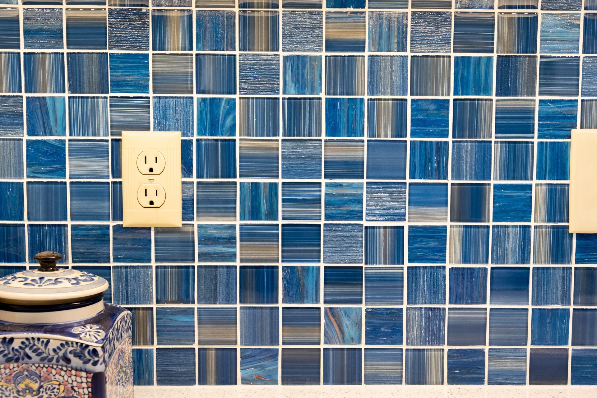 Blue tiled kitchen wall with white electrical outlets and a patterned jar at the lower left