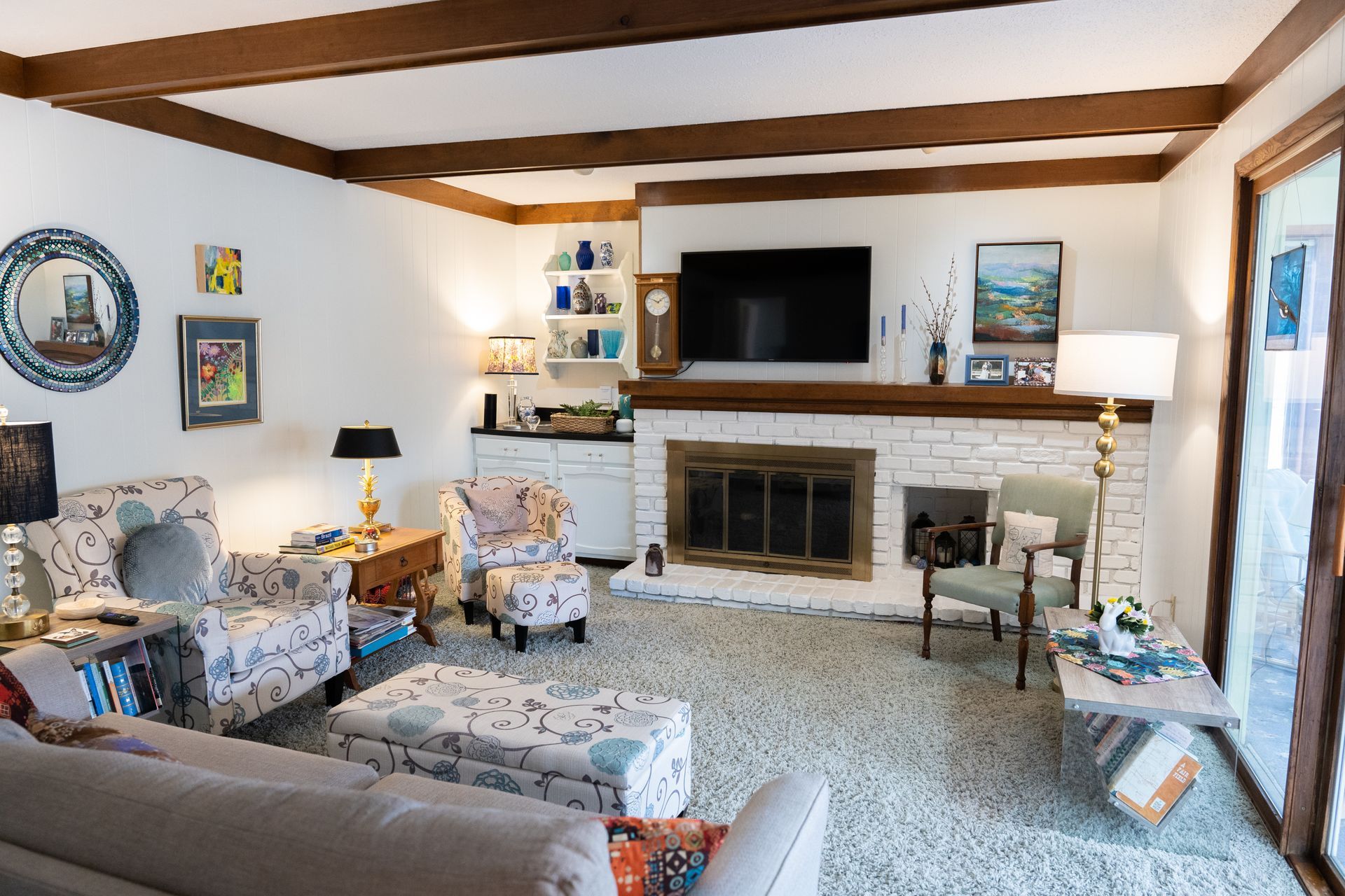 Cozy living room with white walls, wood beams, fireplace, TV, patterned armchairs, and blue accents