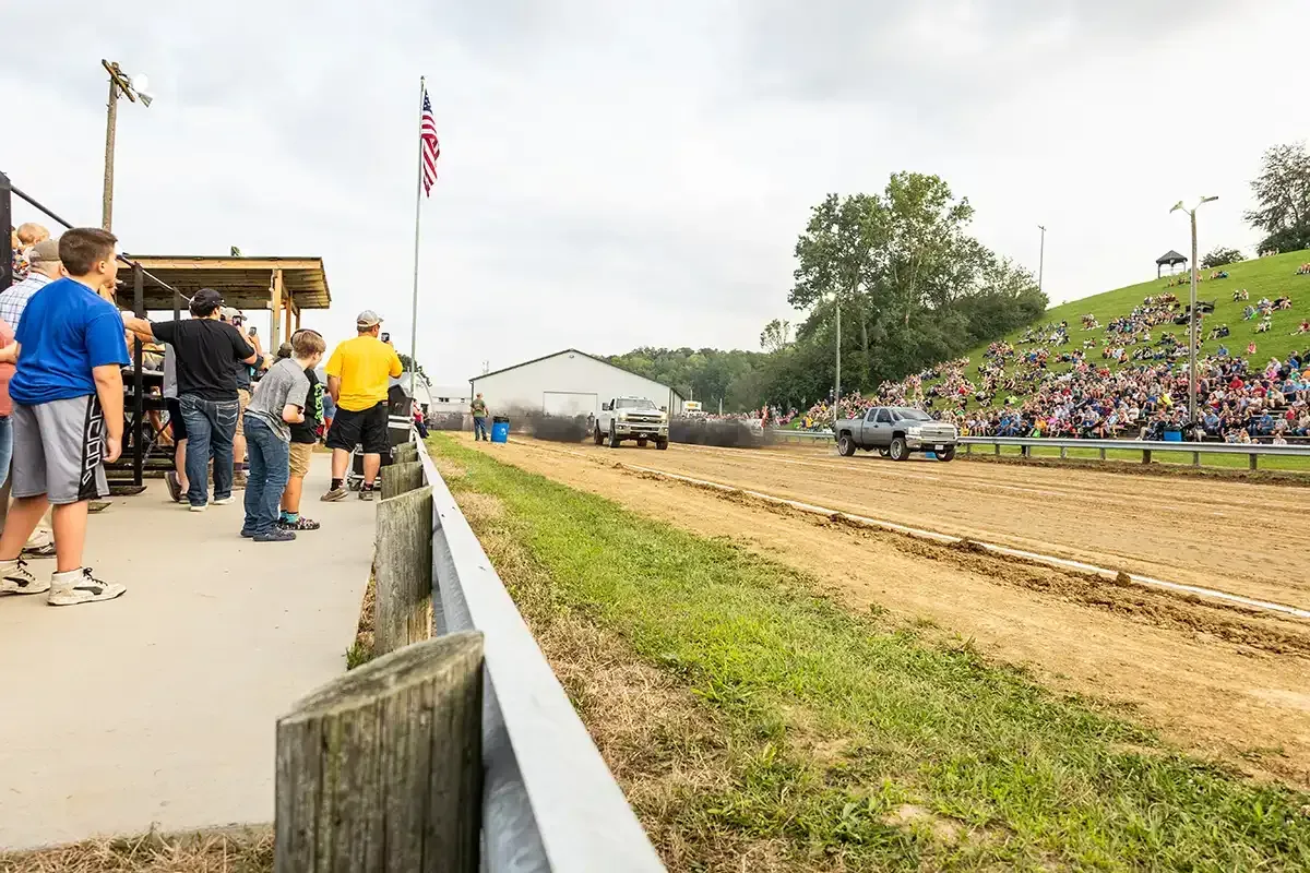 Dirt track fairground with spectators on the left and a crowd on the grassy hillside bleachers.