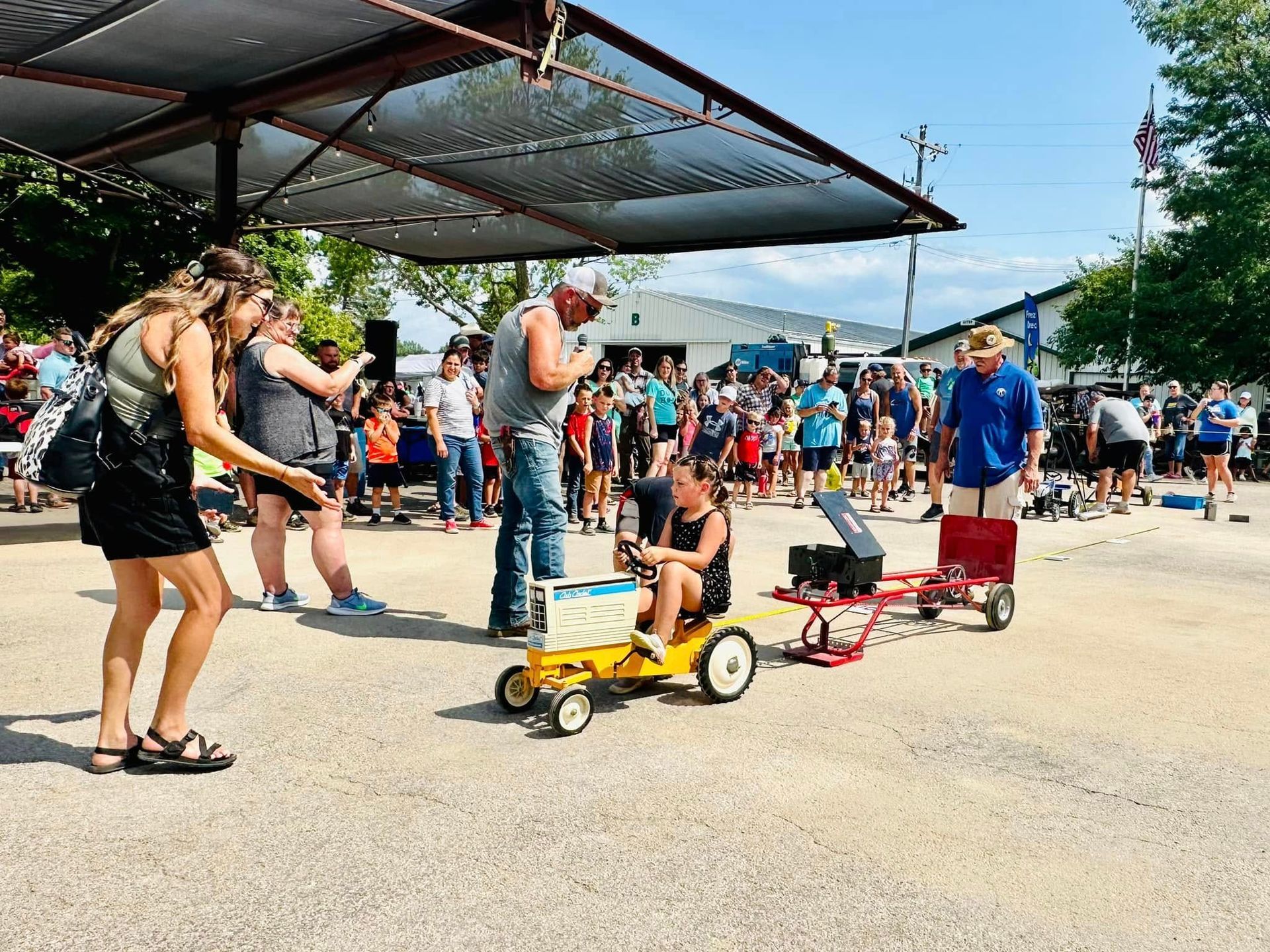 Outdoor event with people watching small children race in pedal cars on a sunny paved lot