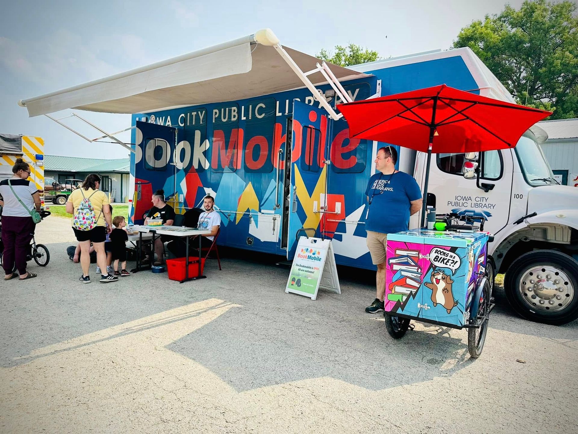 Bookmobile at a library event, with children browsing books and a staff member under a red umbrella.