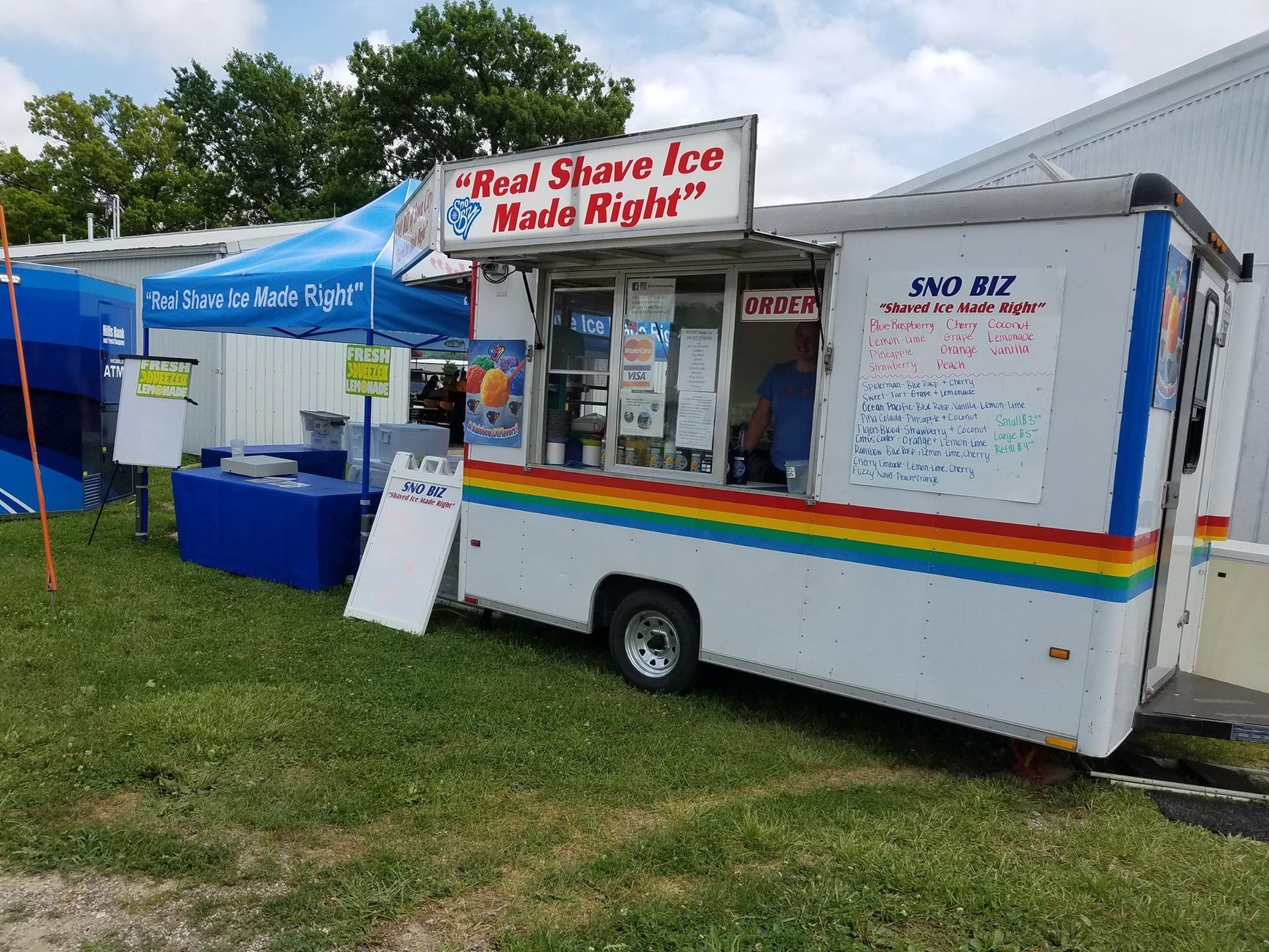 White shaved-ice food trailer with menu boards at a grassy outdoor event, beside a blue canopy