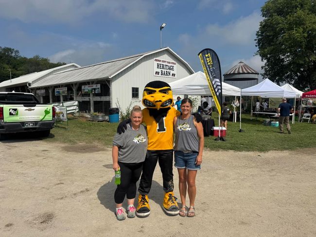 Two women pose with a yellow-and-black mascot outside a white building at an outdoor event.