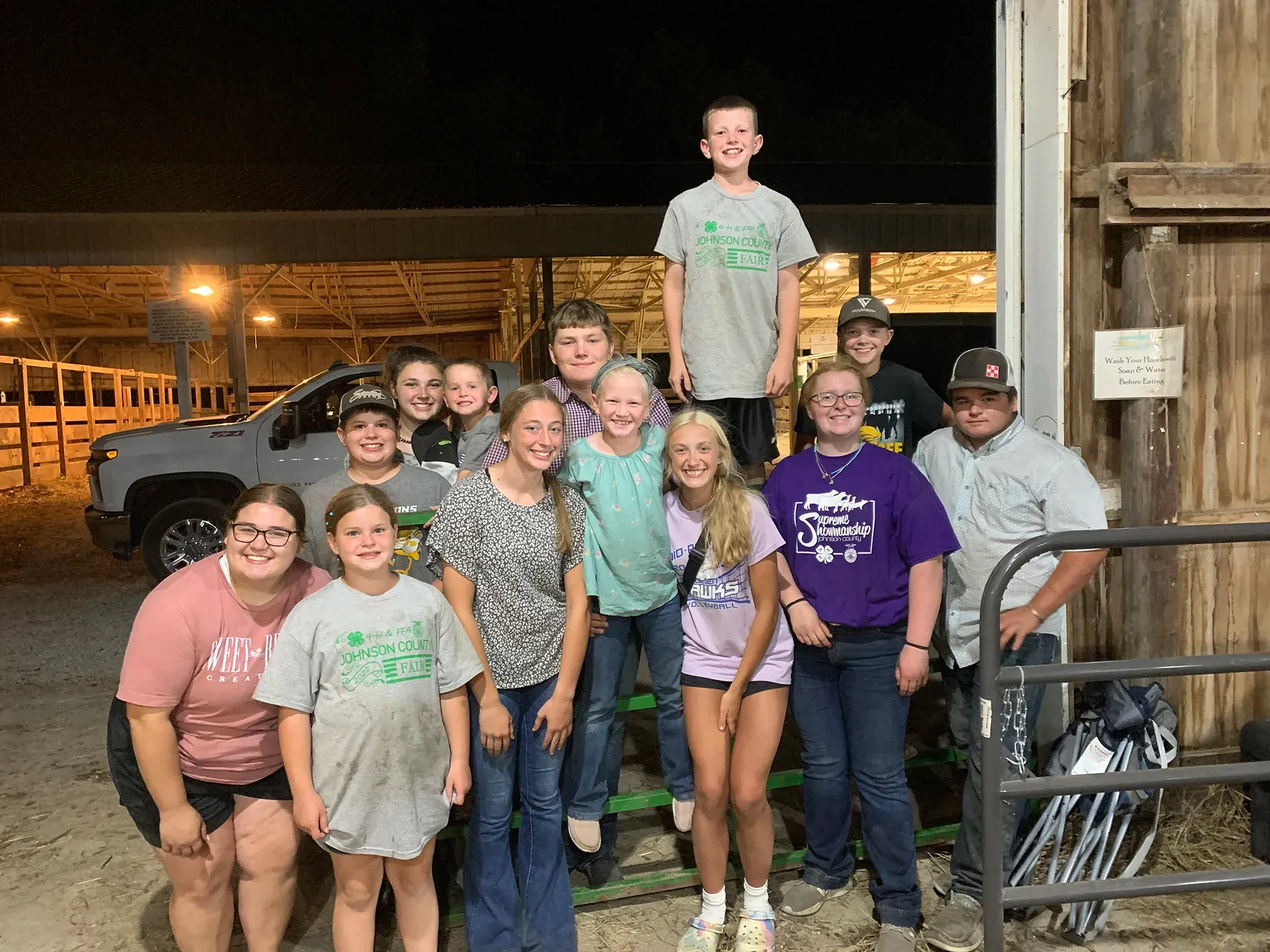 Group of smiling people posing indoors beside a barn wall, with a tall young man centered in back.