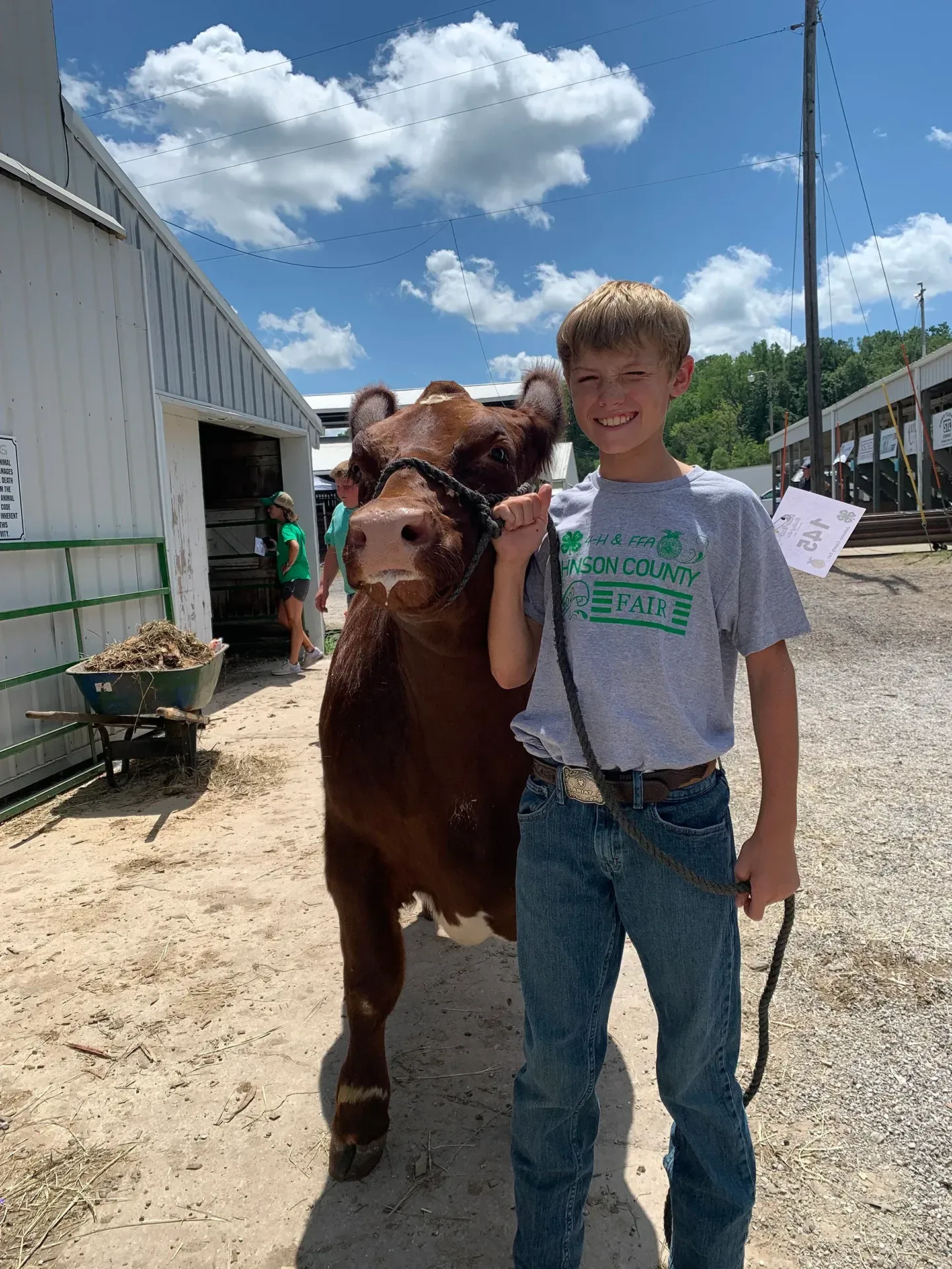 Boy in jeans and gray T-shirt standing with a brown calf outside a barn on a sunny day