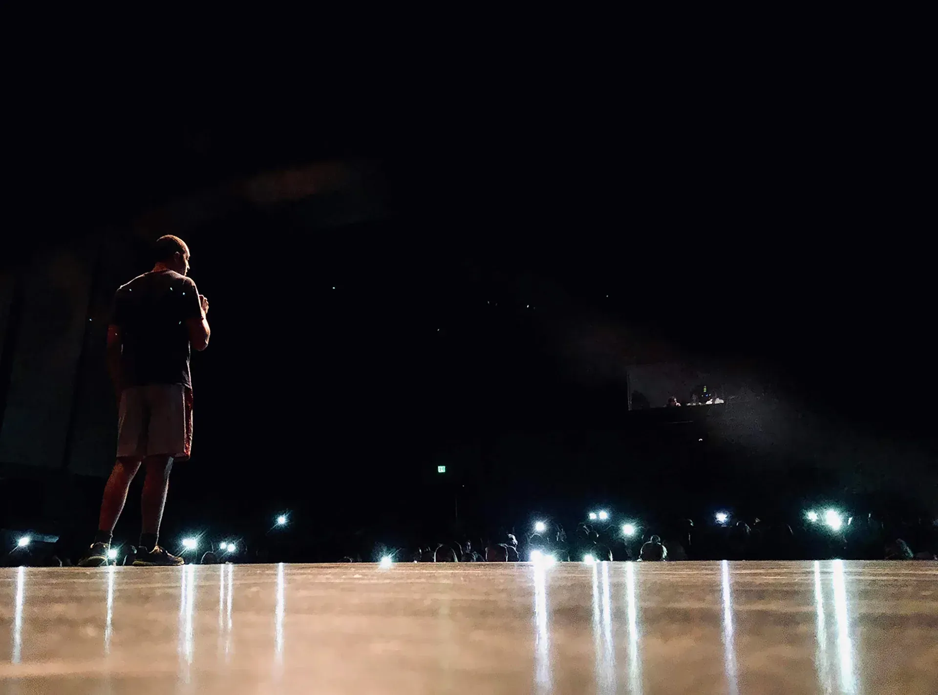Person standing on a stage under spotlights, facing a dark audience with phone lights visible