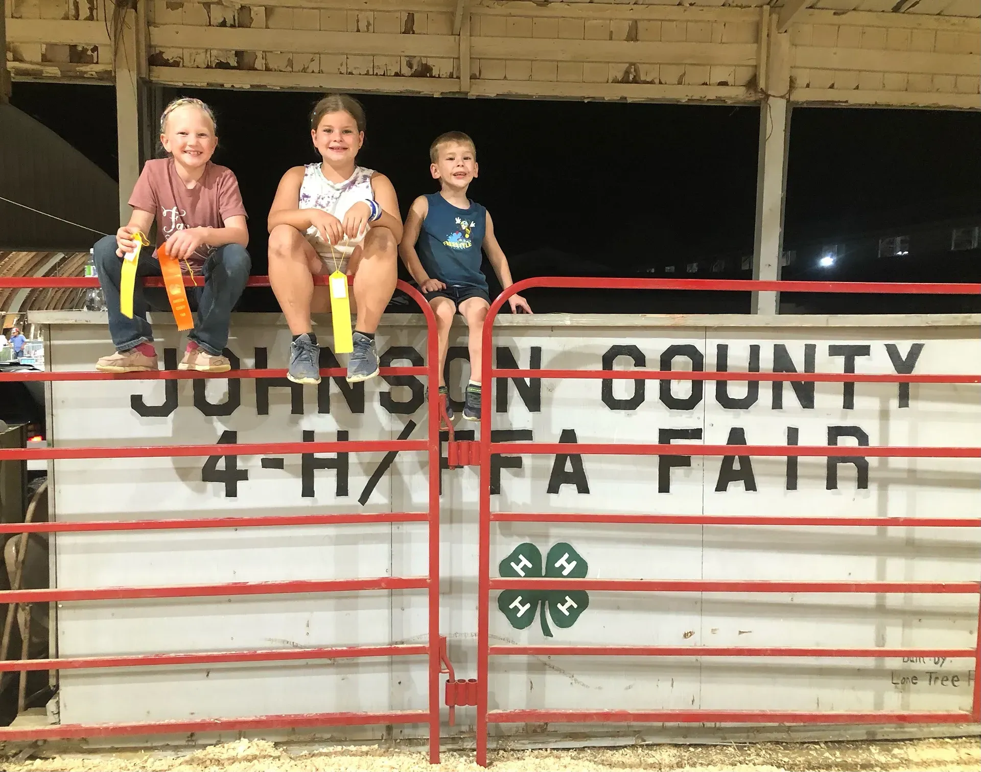 Three children sit on a red gate at the Johnson County 4-H Fair livestock pen.