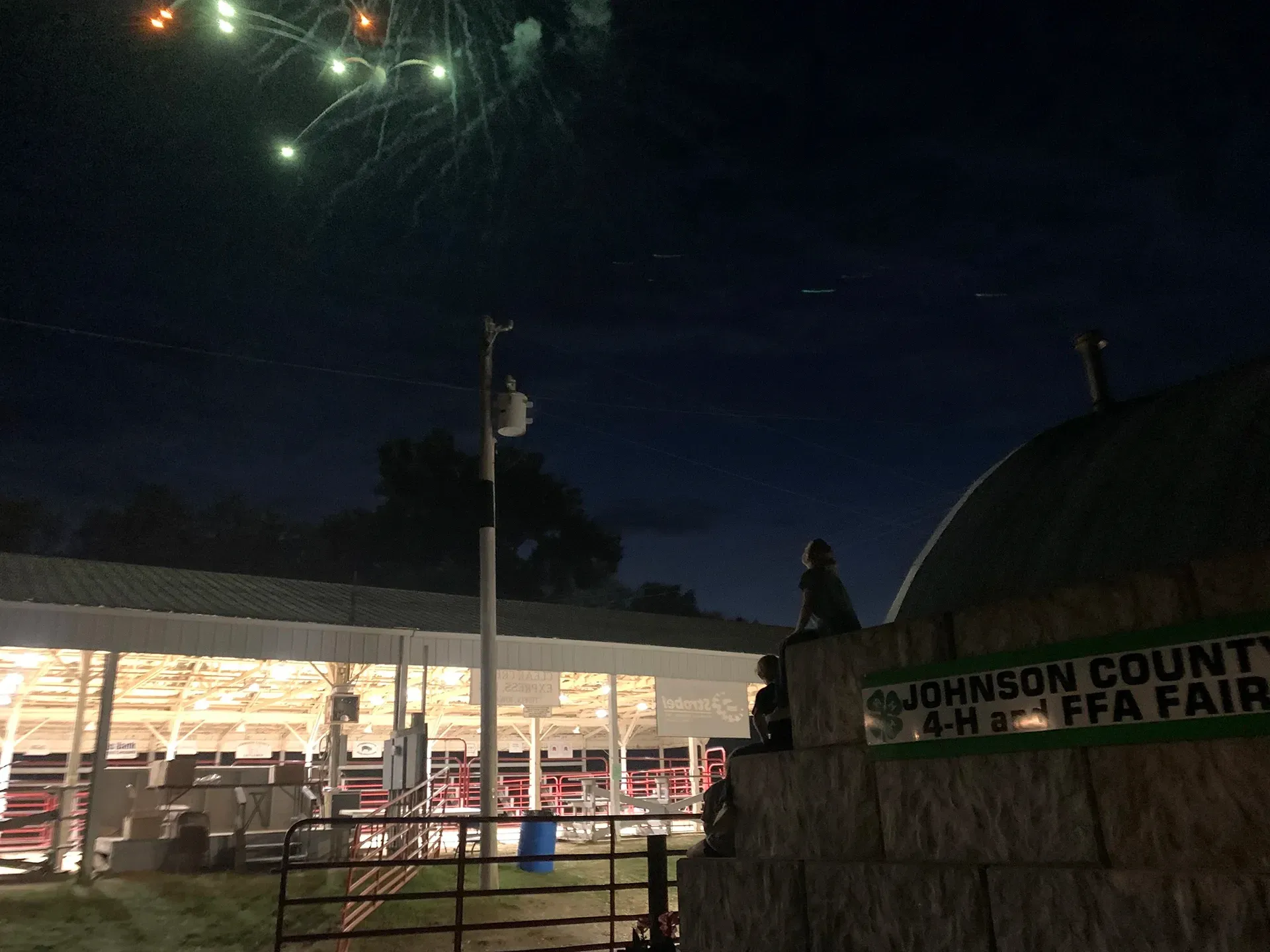 Fireworks over a county fair at night, with a lit pavilion and a large fair sign in the foreground