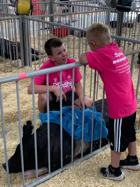Two children in bright pink shirts tending a black pig in a fenced livestock pen