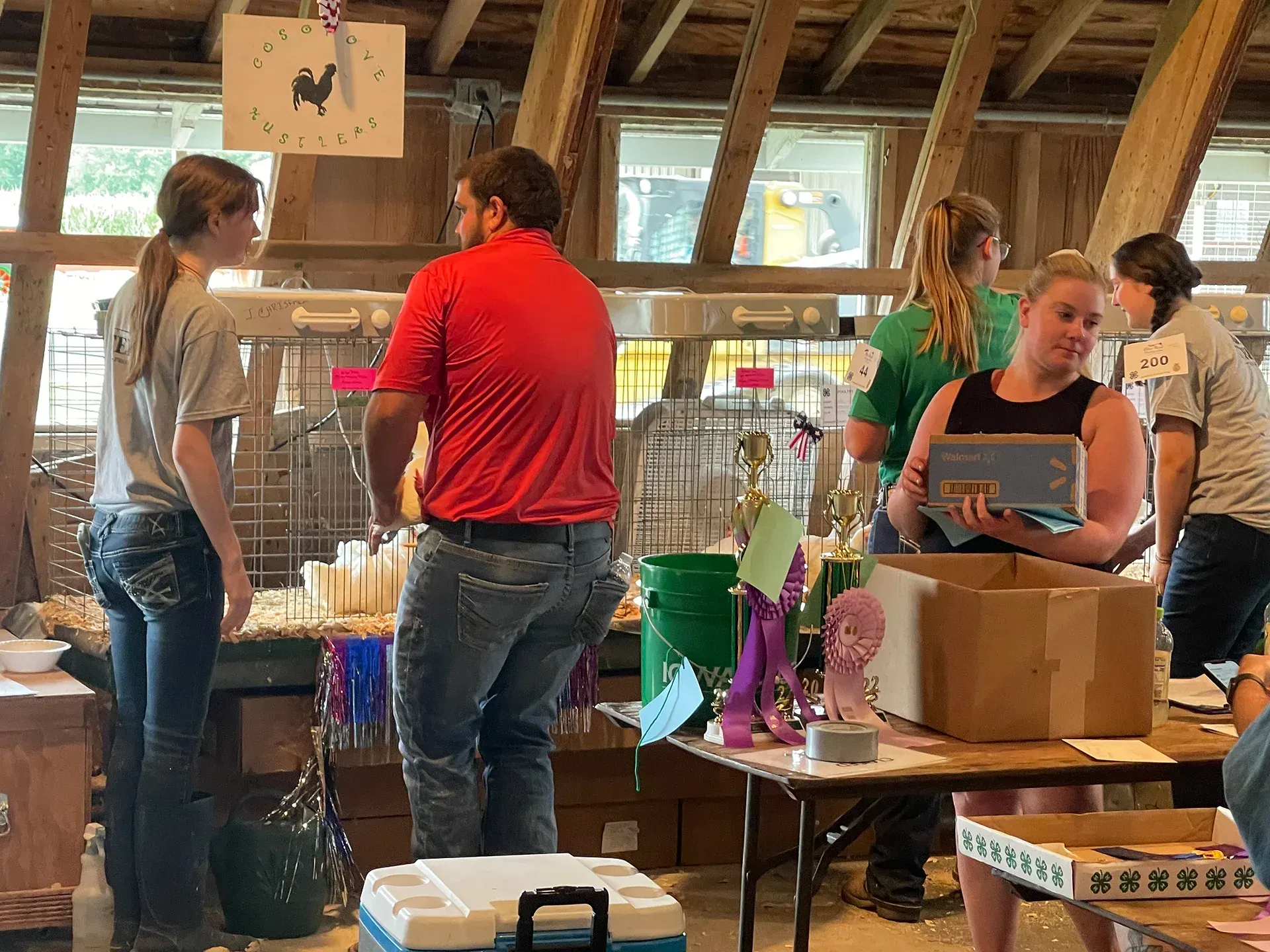 People browsing crafts at a table in a rustic indoor market, with boxes and colorful items displayed.