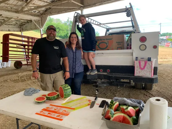 Three people at a farm stand beside a pickup truck, with sliced watermelon and a box of whole watermelons on a table.