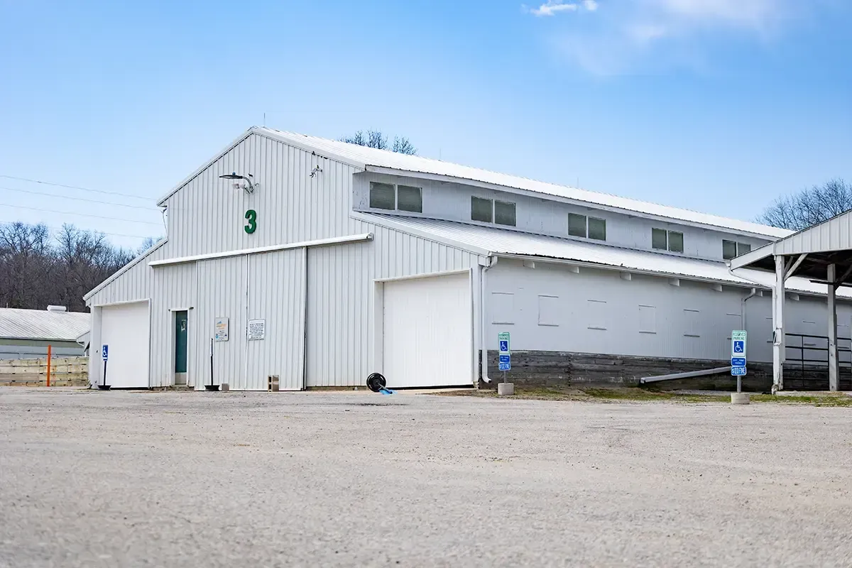 White industrial building with a metal roof and small windows, beside a gravel lot under a blue sky