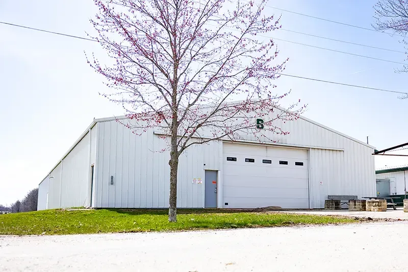 White warehouse with a large garage door, a tree in front, and a grassy lot under a clear sky