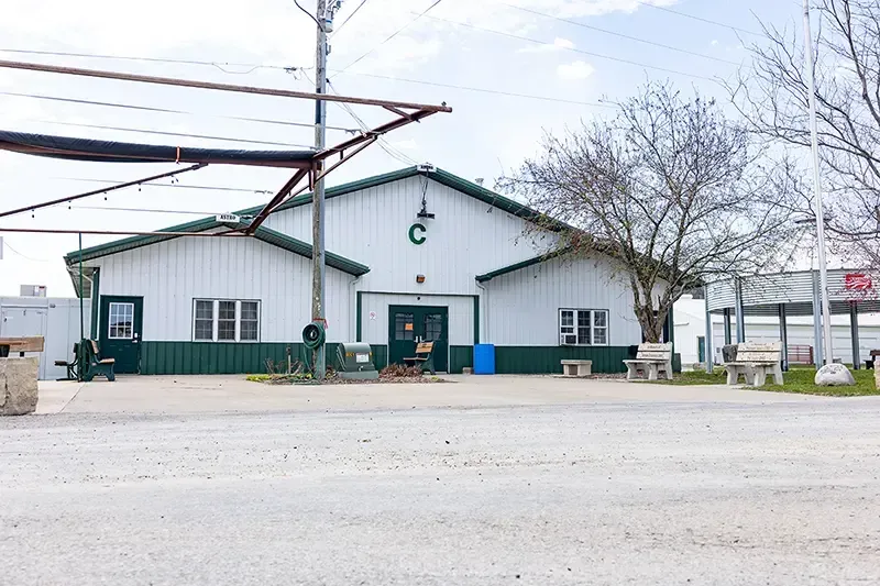 White industrial building with green trim and a large “C,” seen across a gravel lot under bare trees.