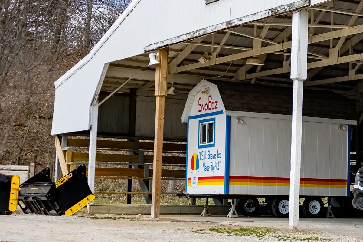 Small white trailer with blue trim parked under a barn-like shelter beside a black-and-yellow tractor attachment.