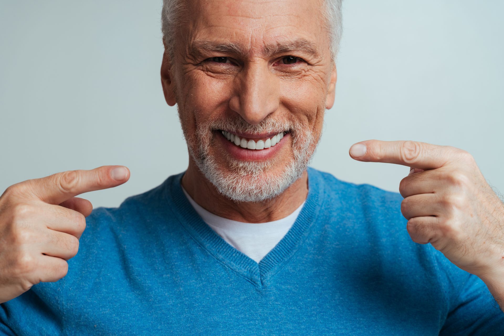 Man with gray beard and white teeth smiles, pointing at his teeth with both index fingers.