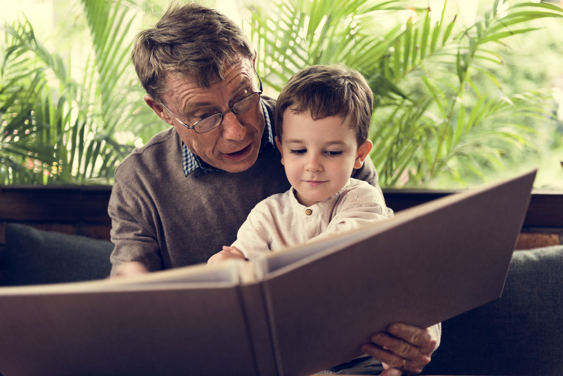 Older man reading a large book with a young child on a couch, with green plants in the background
