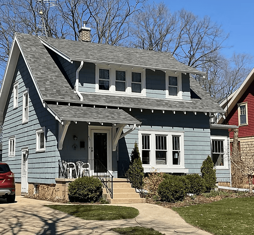 Gray two-story house with front porch, dormer windows, and a small front yard on a sunny day