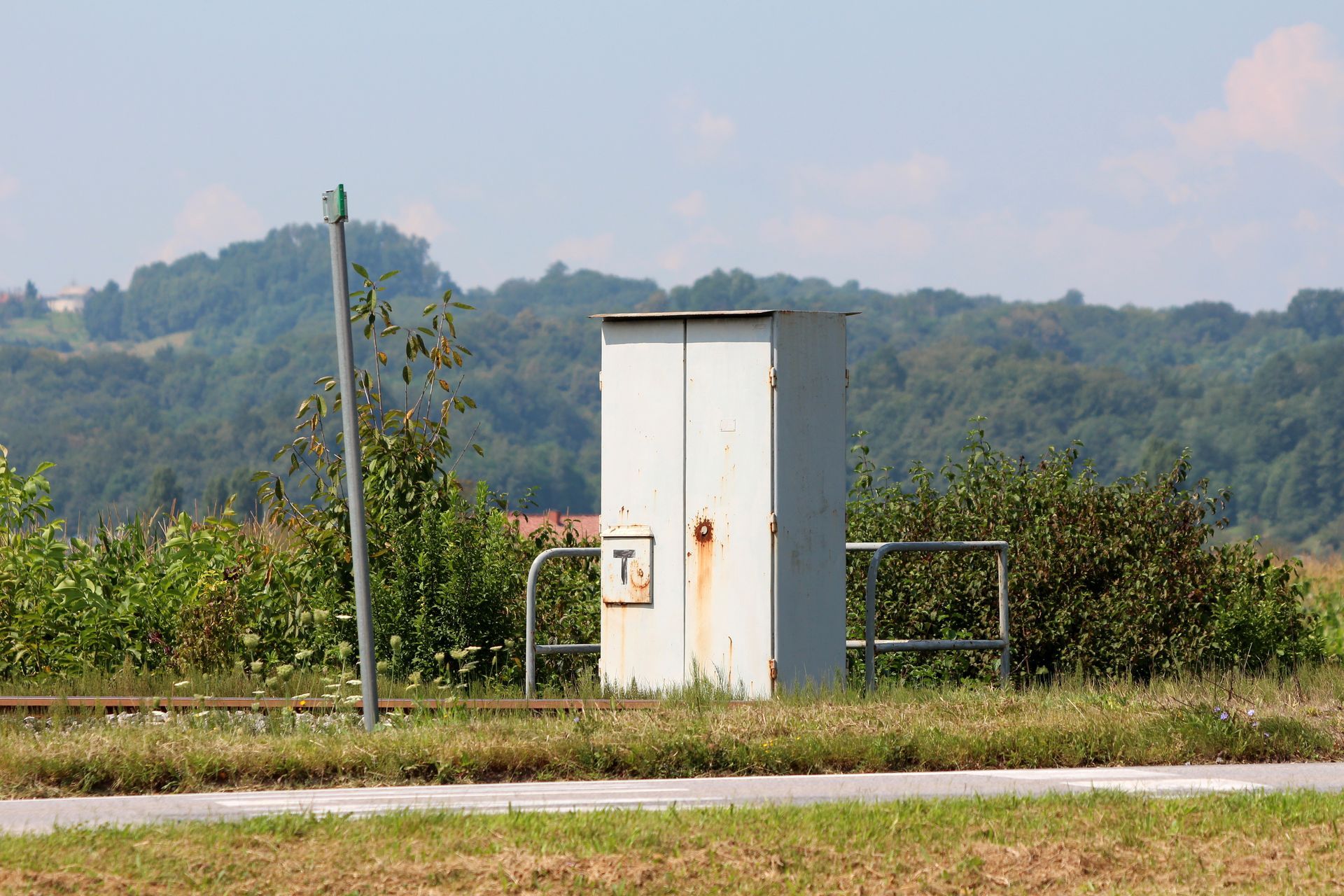 Small white utility cabinet beside a road, with green fields and wooded hills in the background.