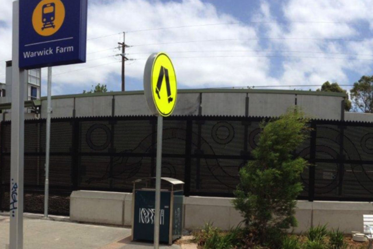 Warwick Farm station entrance with yellow pedestrian sign, fence, and trees under a cloudy sky