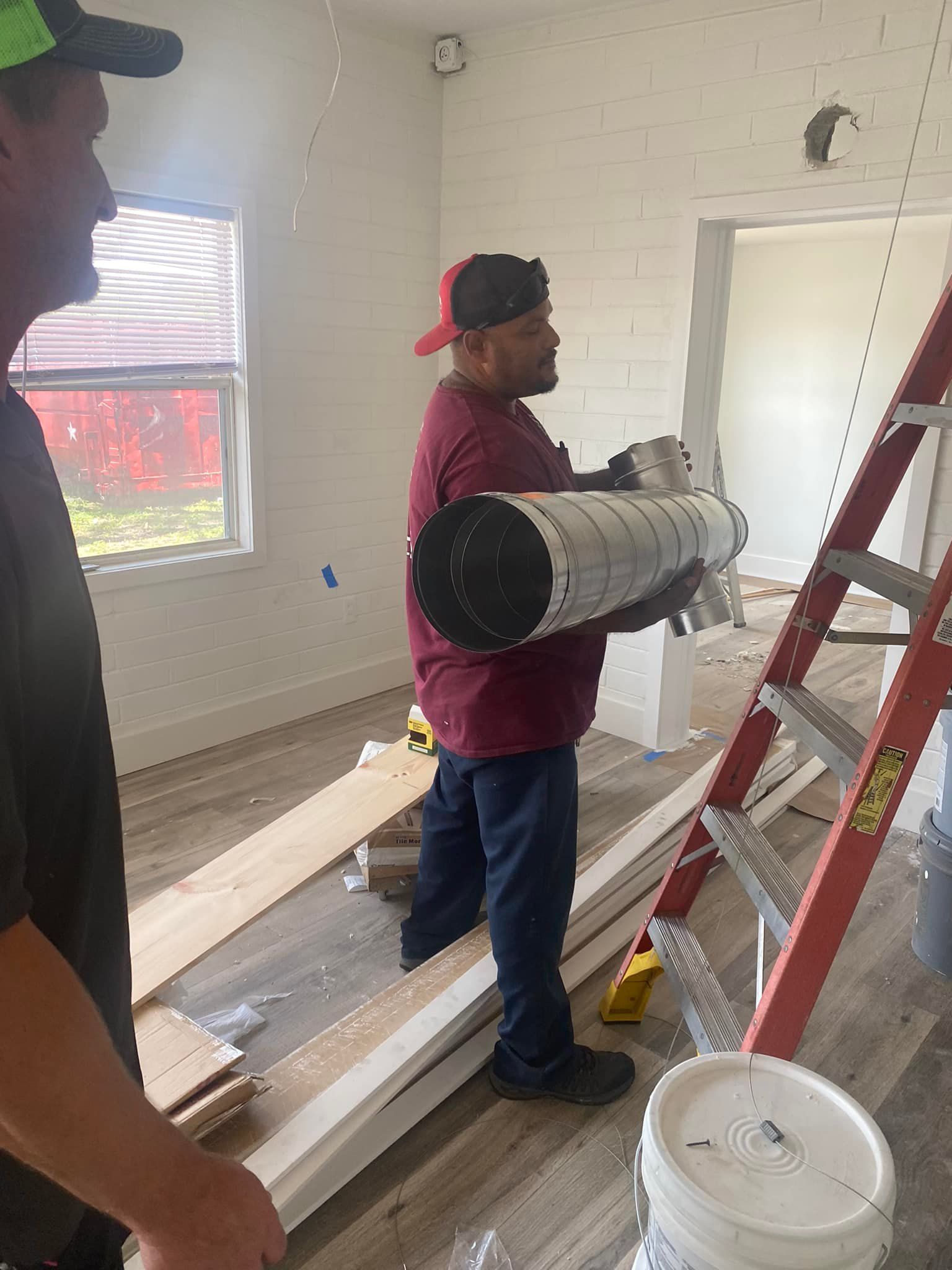 A man is carrying a roll of carpet in a room next to a ladder.