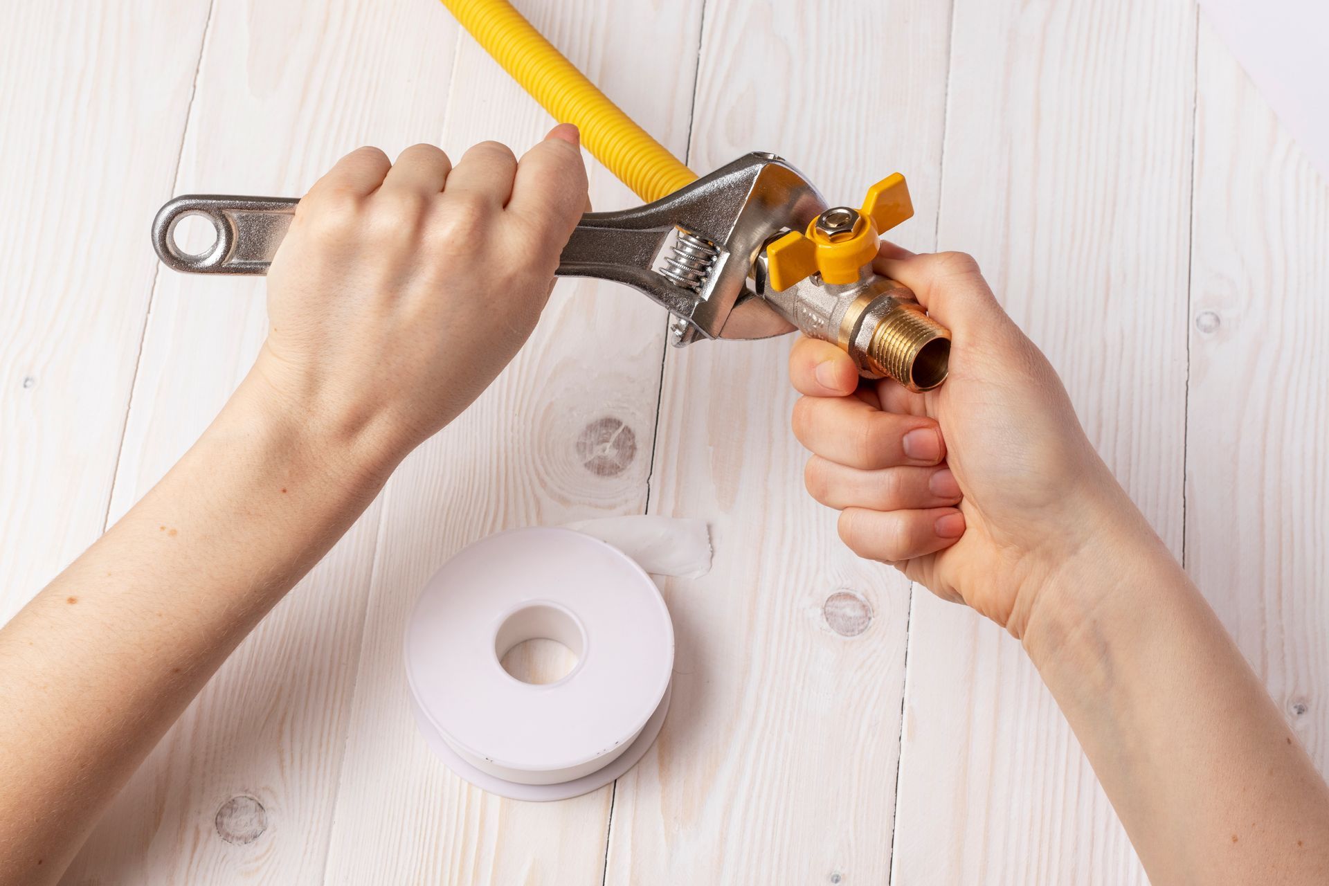 Hands tightening a gas line fitting with a wrench, yellow pipe visible, white thread tape nearby.