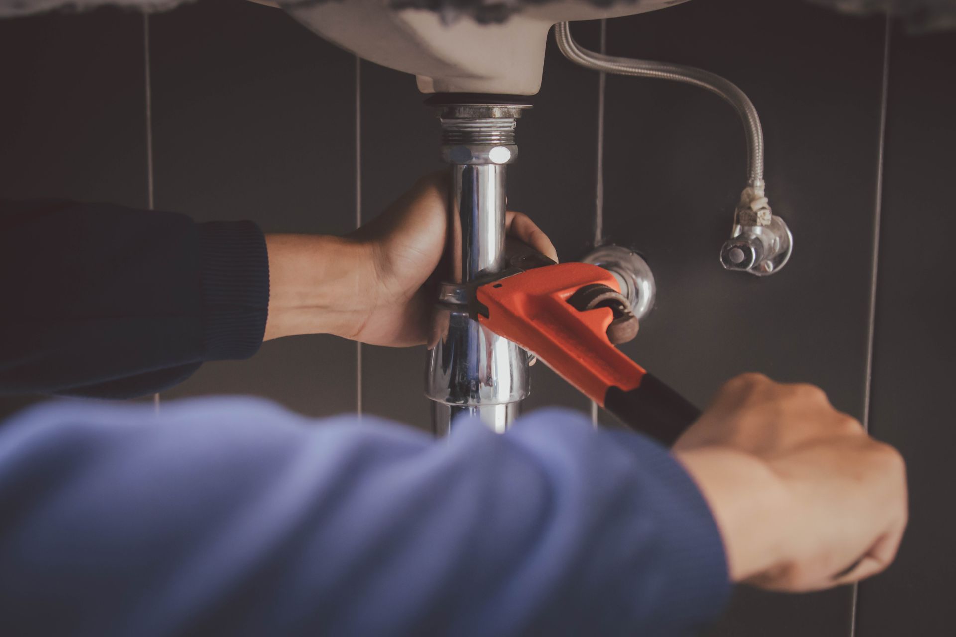 A plumber in a blue shirt uses a wrench on a sink's drain pipes.