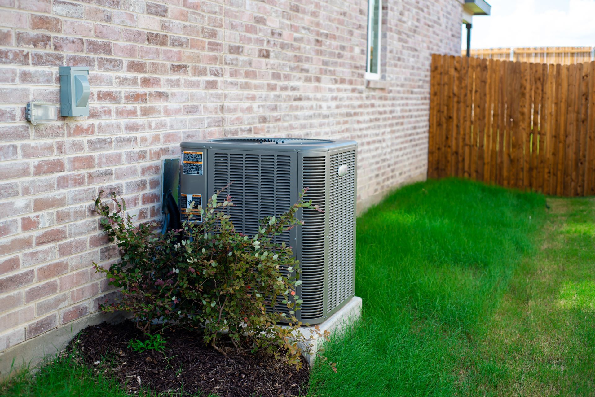 Air conditioning unit next to a brick building and a wooden fence, surrounded by grass and mulch.