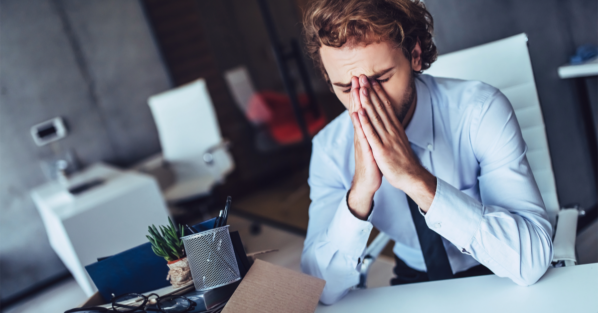 A man is sitting at a desk with his hands on his face, stressed as he may be getting fired