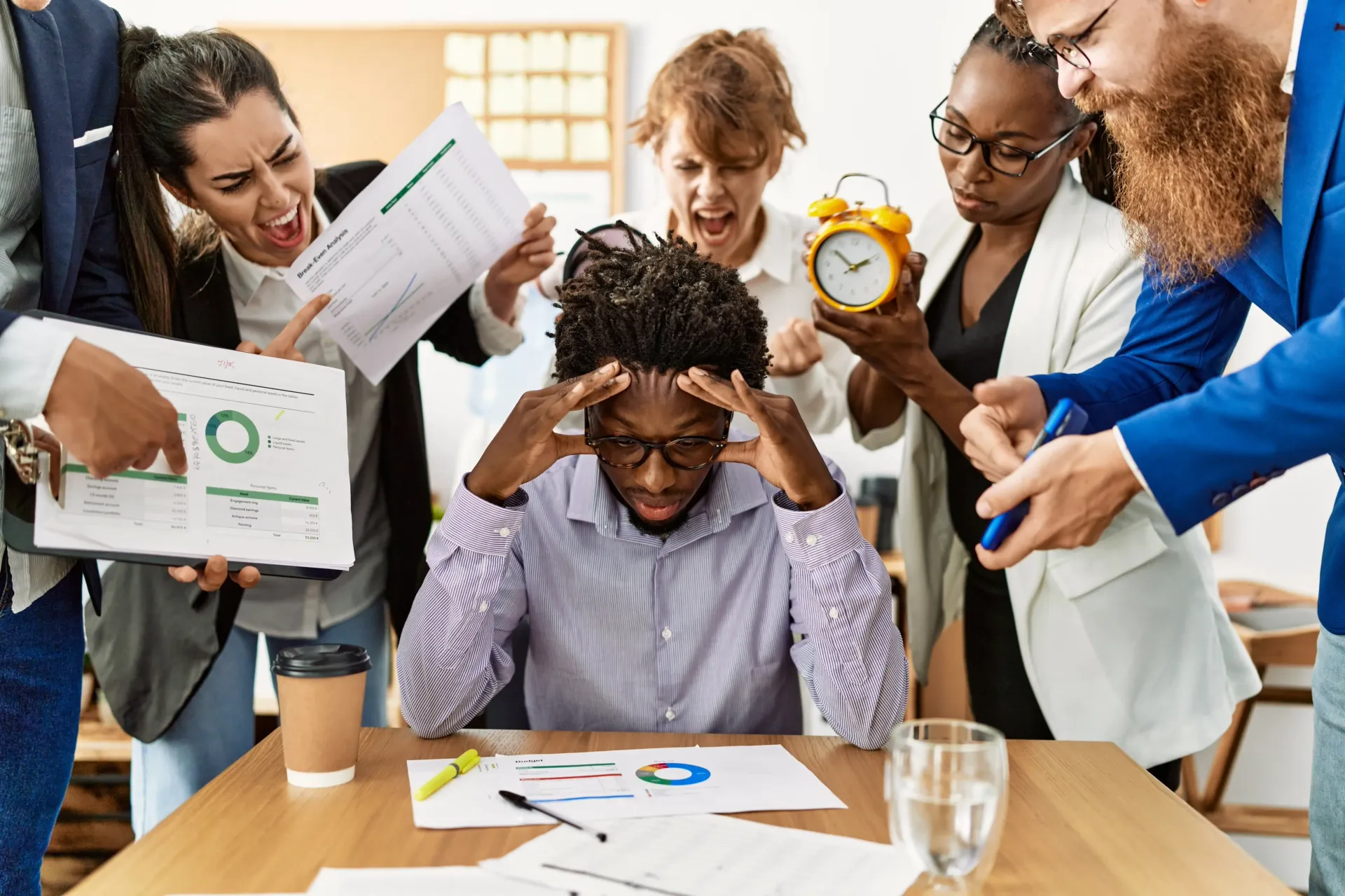 A stressed person surrounded by colleagues pointing, yelling, and holding an alarm clock in an office setting.
