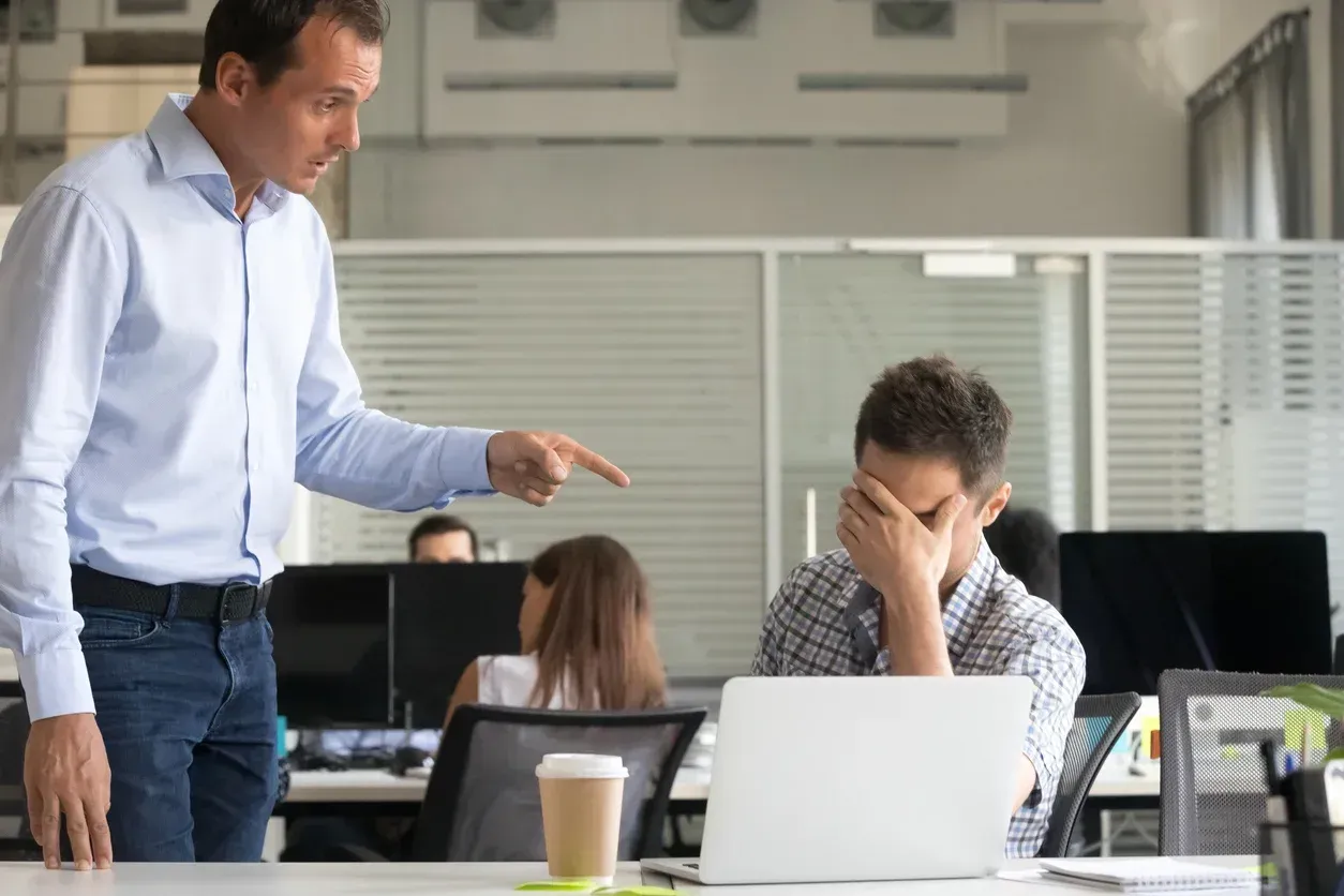 Angry looking man points at another man with his head in his hands at an office desk.
