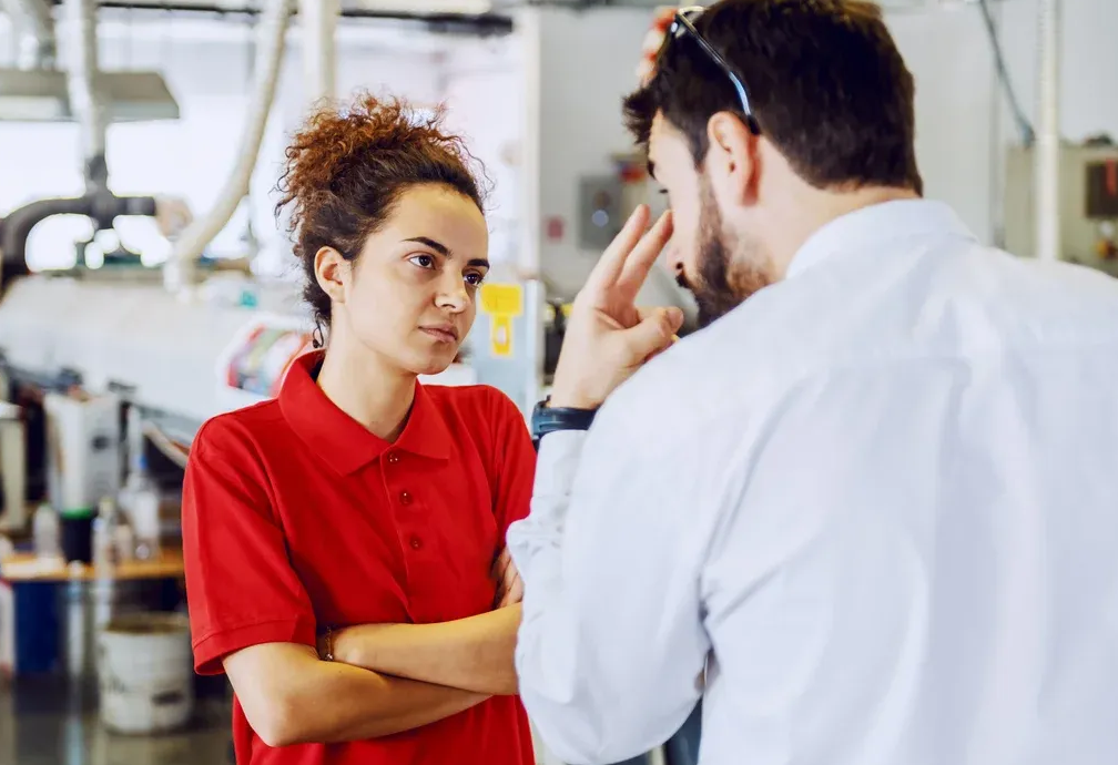 Woman in red shirt and man in white shirt appear to argue in a factory setting.