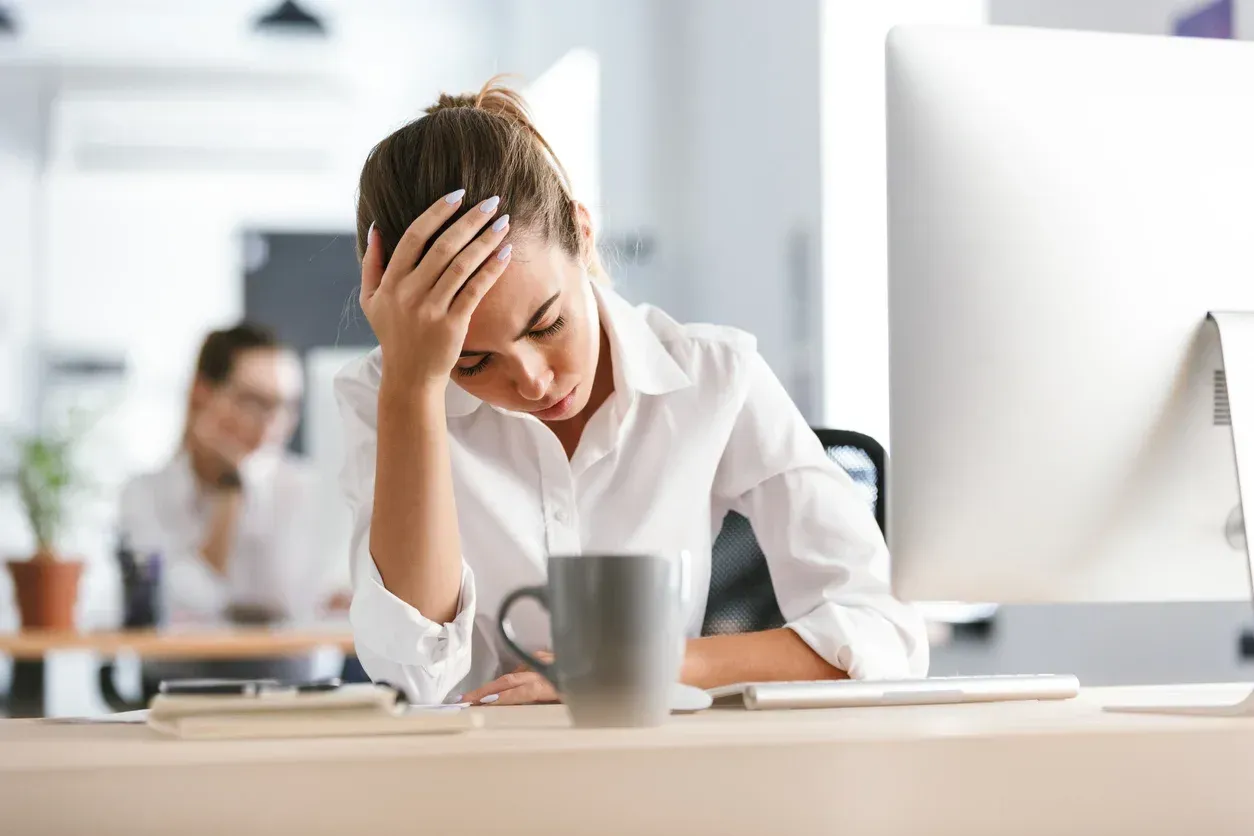 Woman with hand on forehead, looking down at desk, in office setting.