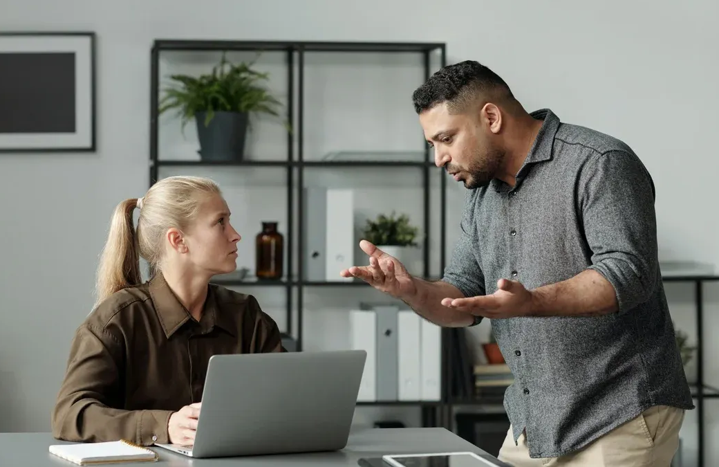Man gesturing at woman, both looking at each other near a laptop in an office setting.