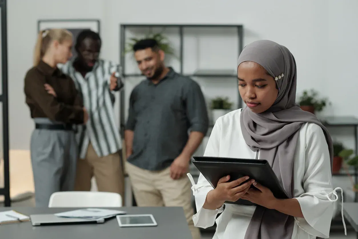 Woman in hijab looks at tablet while three coworkers talk in background.