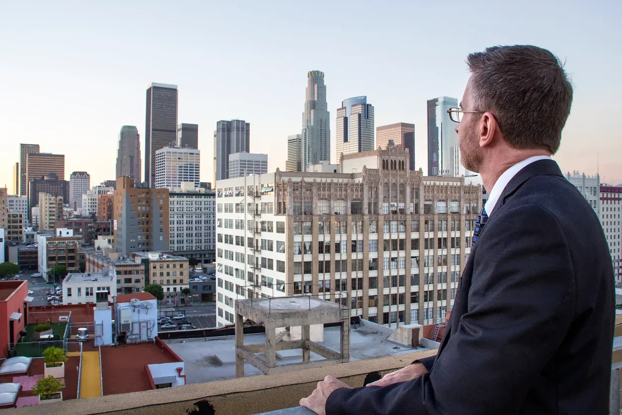 A lawyer in a suit looks at los angeles skyline from rooftop at dusk.