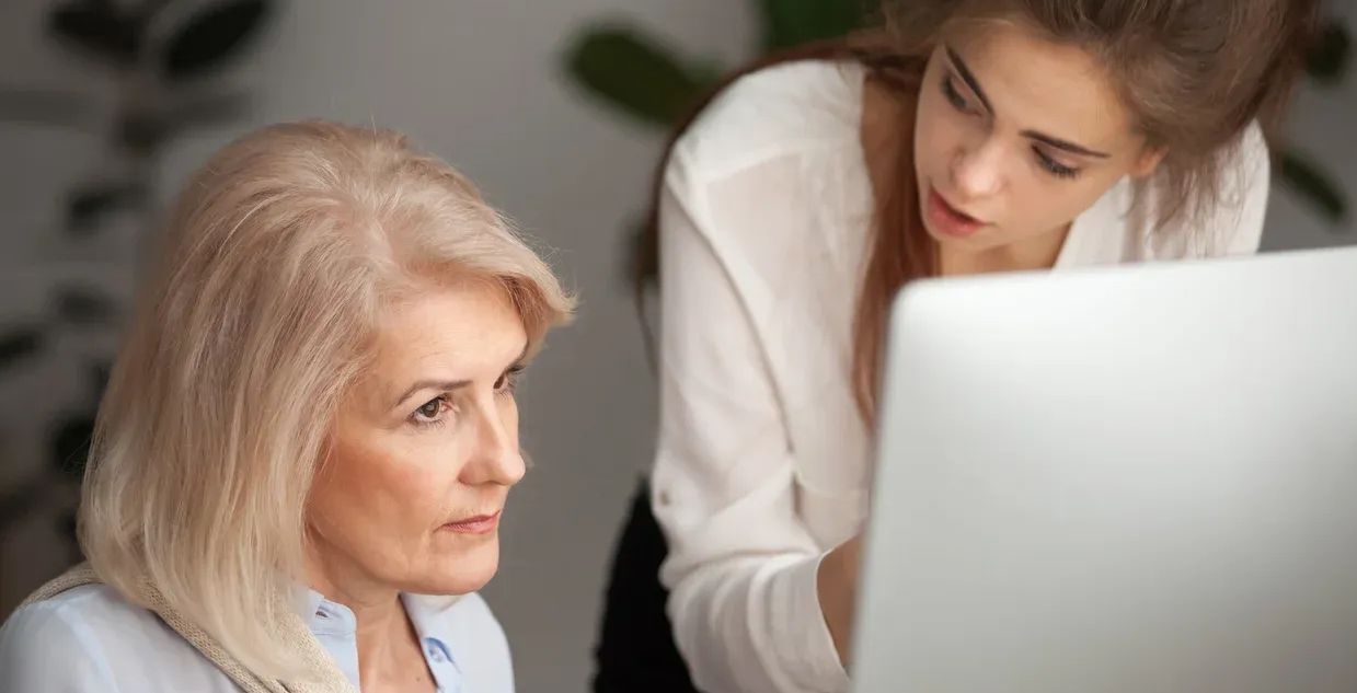 older woman looks at a computer while a younger woman appears to talk down to her
