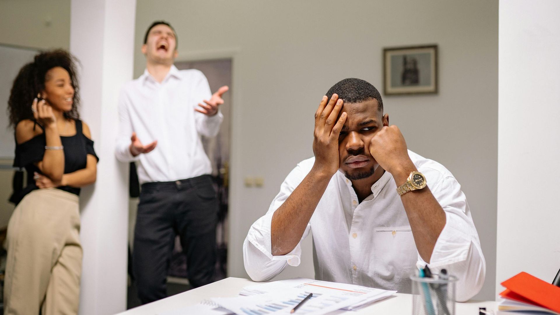 A man is sitting at a desk with his hands on his head, and a man is standing behind him.
