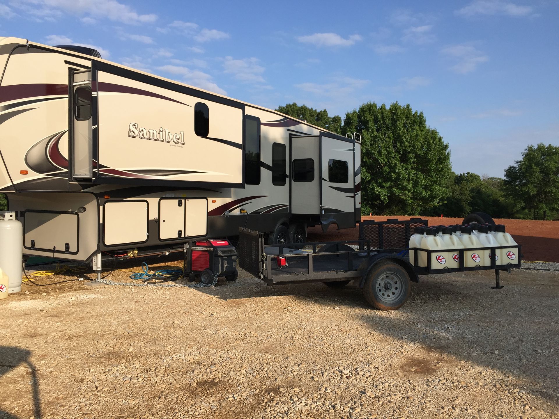 Large beige fifth-wheel RV parked on gravel beside a small flatbed trailer under a clear sky.