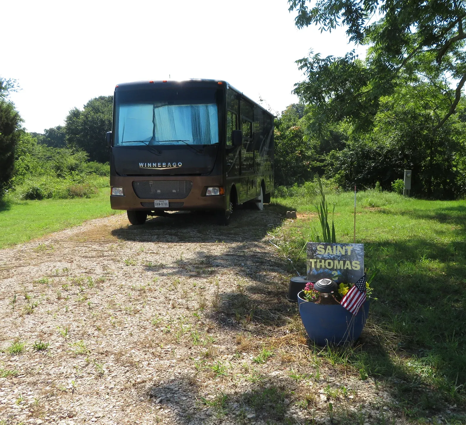 Brown motorhome parked on a gravel driveway beside grass and trees, with a blue planter in the foreground.