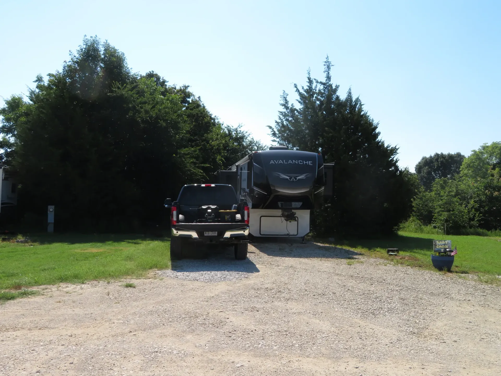 Truck and RV parked on a gravel driveway beside trees under a bright sky