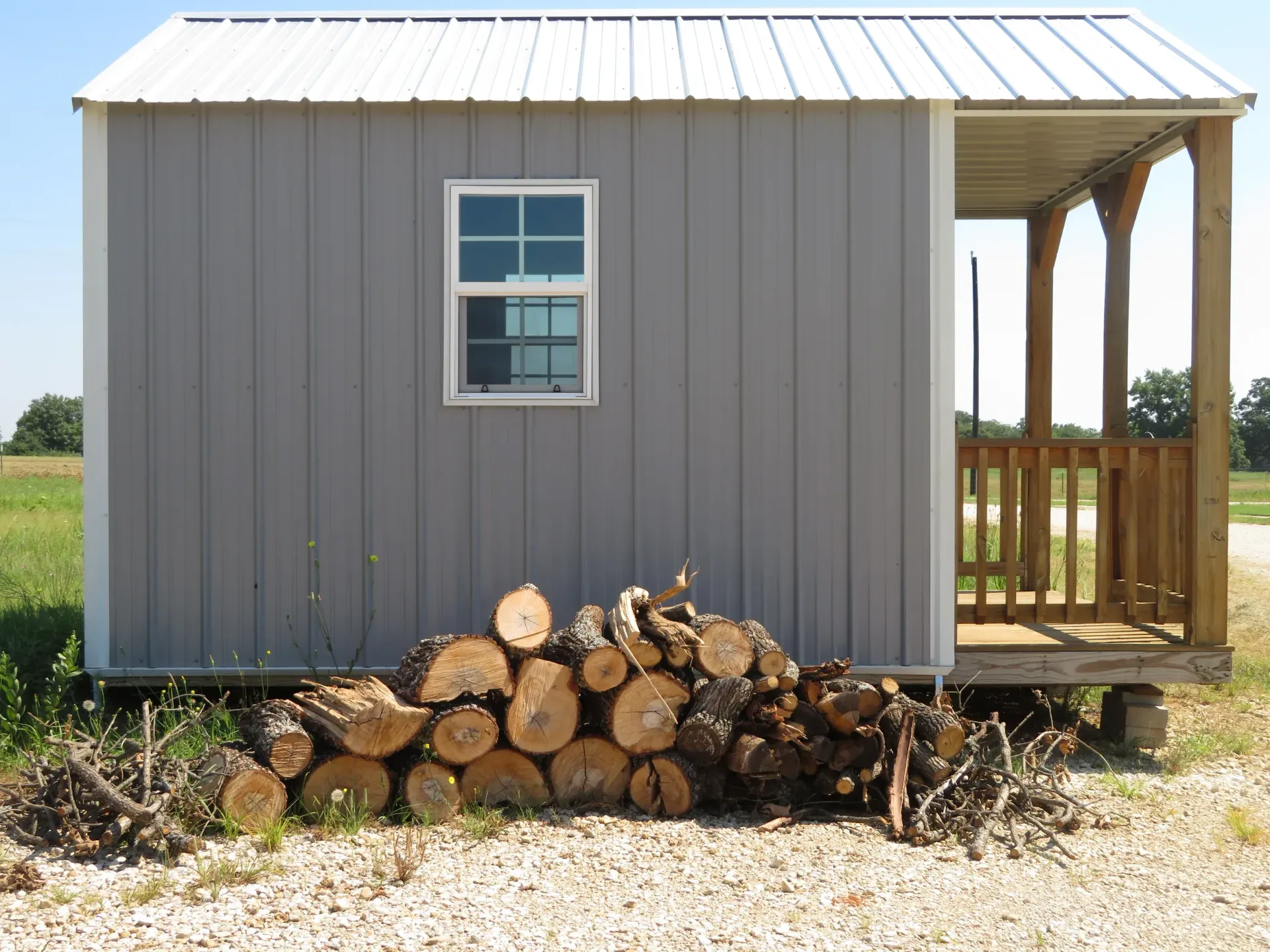 Gray shed with a porch and stacked firewood along the base in a grassy field