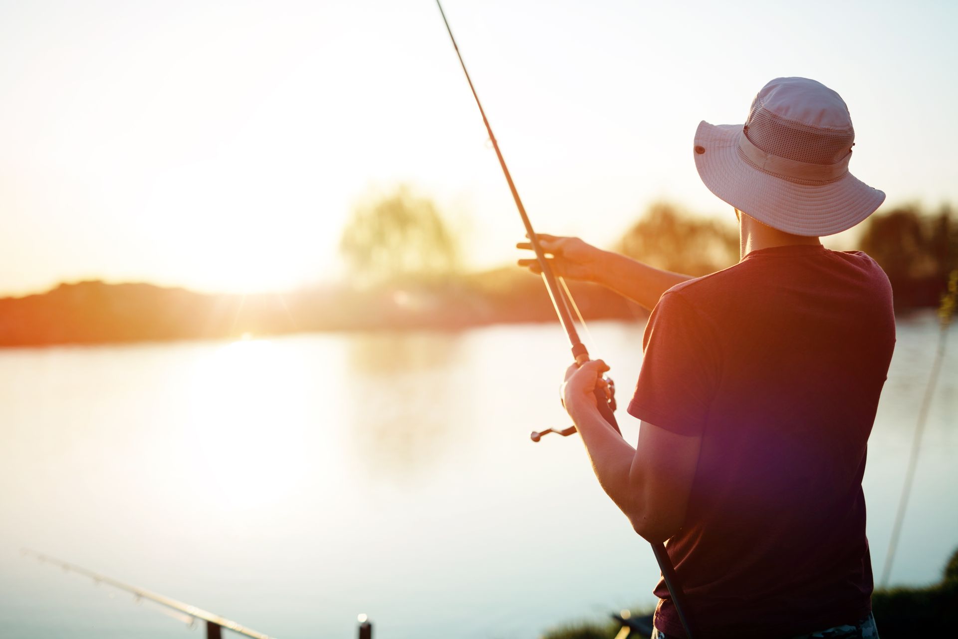 Person fishing at sunset by a calm lake, backlit by warm golden light.