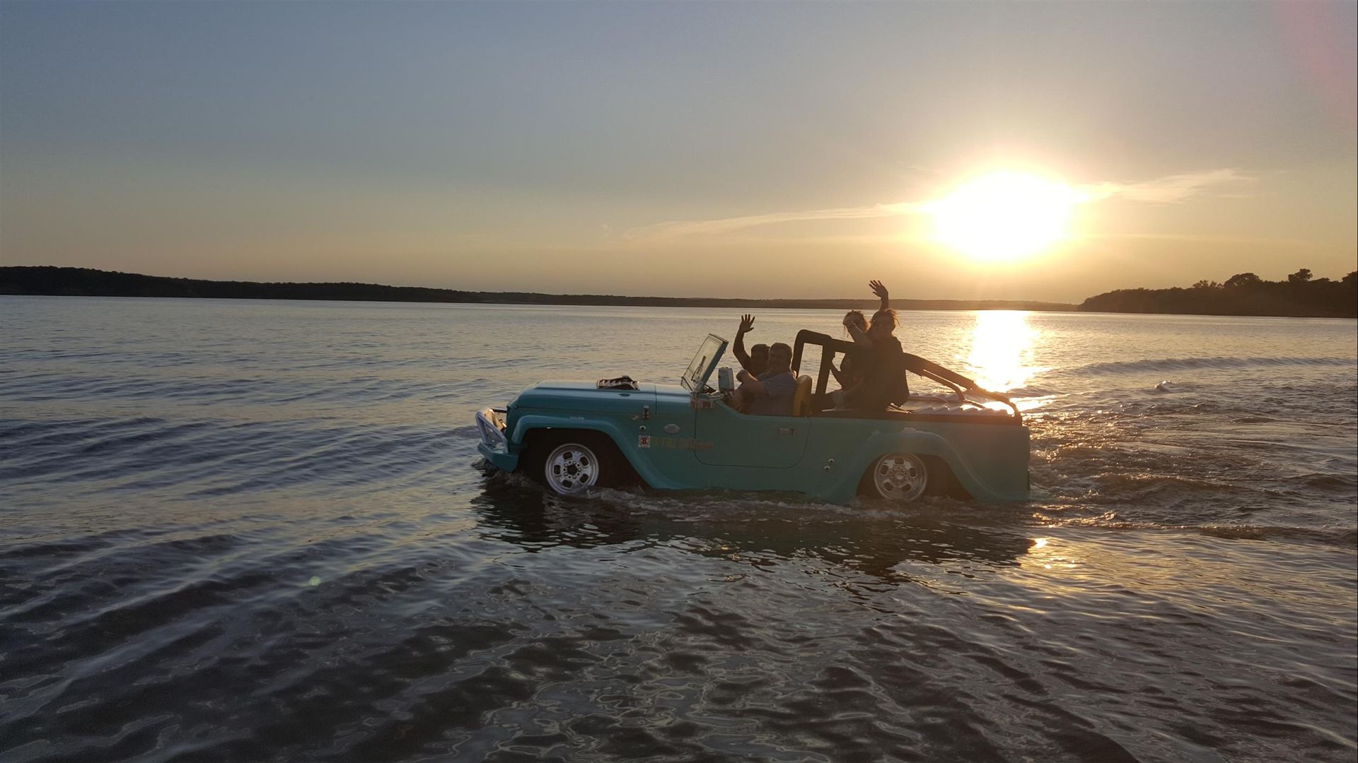 Blue Jeep driving through shallow water at sunset, with people standing in back and sun reflecting on the surface