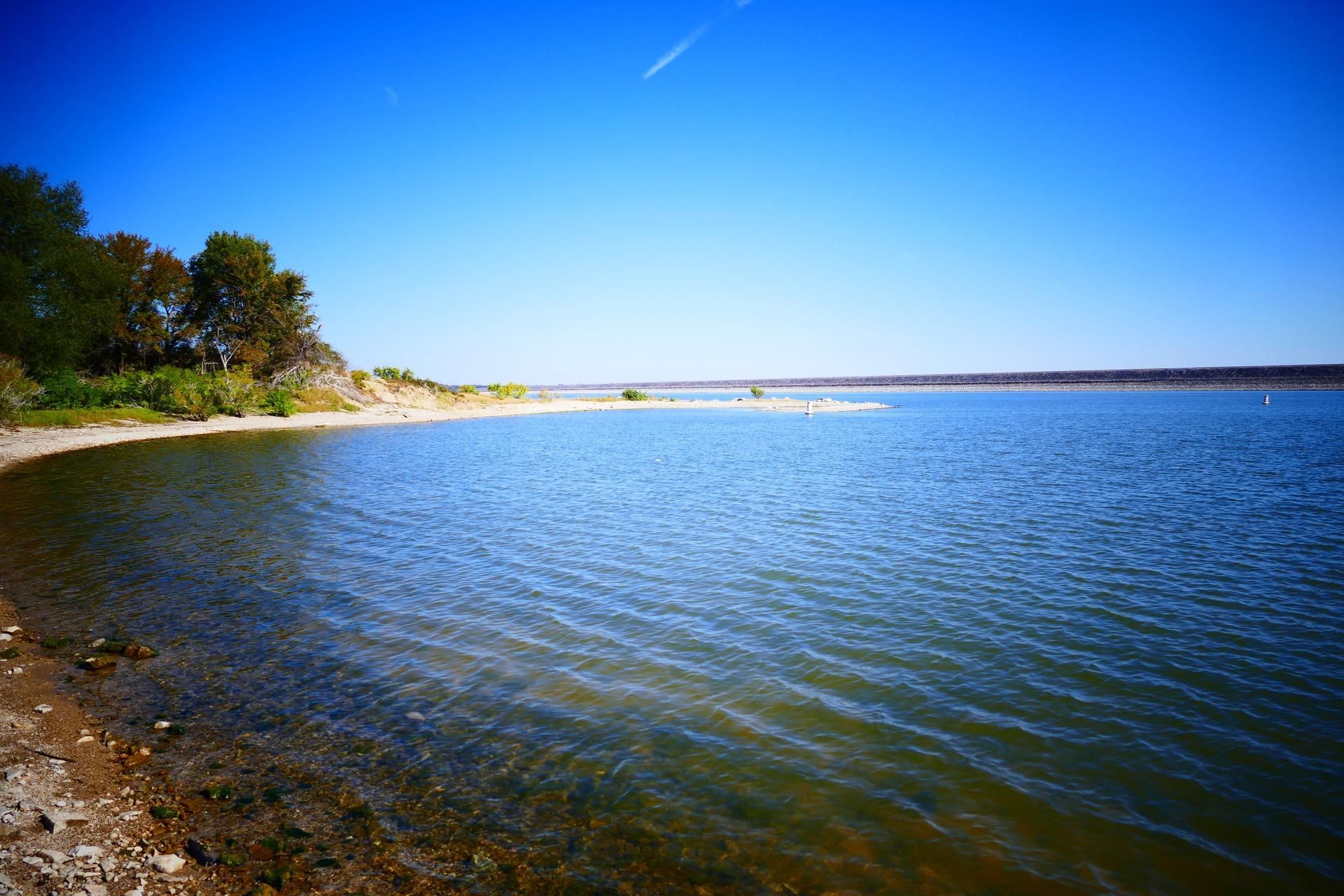 Calm blue bay with rocky shoreline, sandy beach, and trees under a clear sky