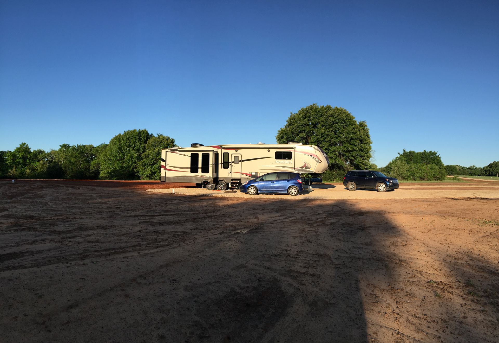 RV parked on a dirt lot with two cars under a clear blue sky