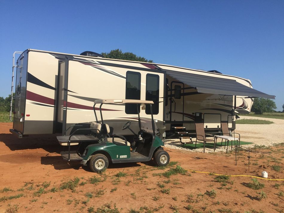 Beige RV with slide-outs parked on a dirt lot under a clear blue sky, with a golf cart hitched in front