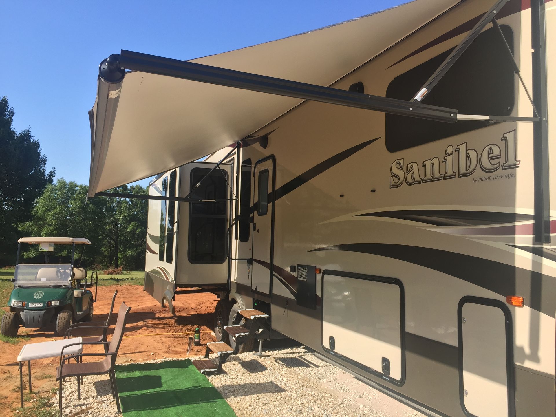 RVs with a beige awning extended over a sandy campsite, with a golf cart and chairs nearby